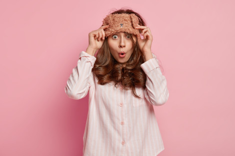 Astonished dark haired young cute European woman has eyemask on forehead, wears pajams, wonders to have dreadful dreams, poses against pink background. People, bed time and surprisement concept.